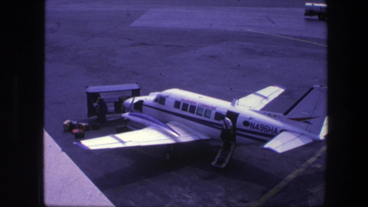 1975 Copenhagen home-movie frame showing a small white commuter plane marked N496HA on the tarmac with ground equipment nearby.