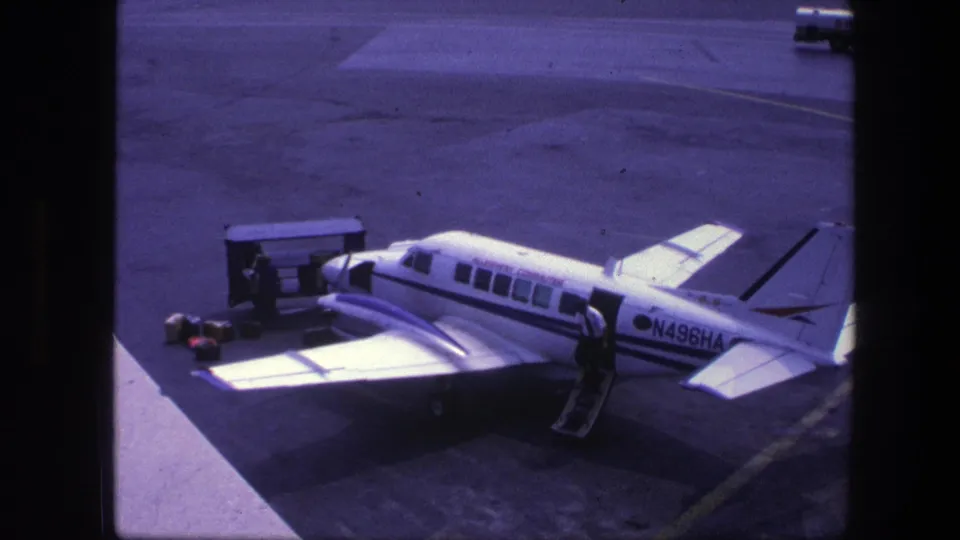 1975 Copenhagen home-movie frame showing a small white commuter plane marked N496HA on the tarmac with ground equipment nearby.