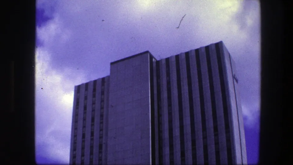 1975 Copenhagen home-movie frame with a low-angle view of a tall modern high-rise facade beneath a cloudy sky.