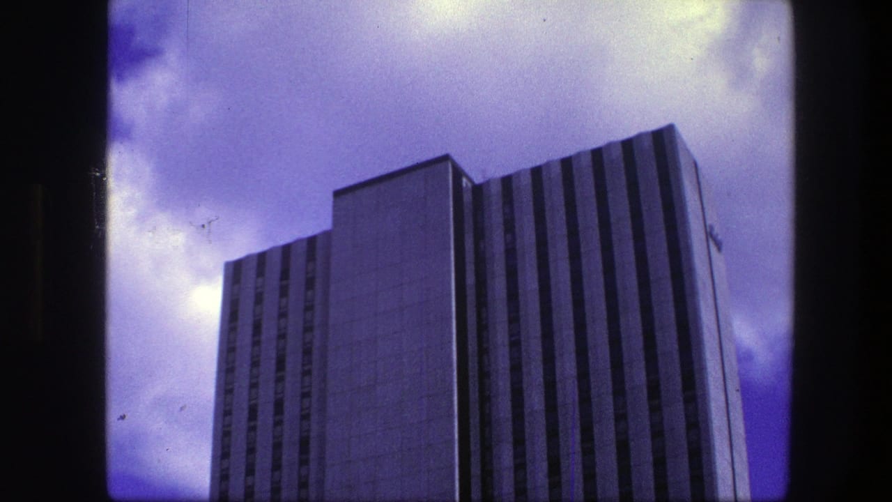 1975 Copenhagen 8mm frame showing a tall modern office building from below, with vertical facade lines and cloudy sky.