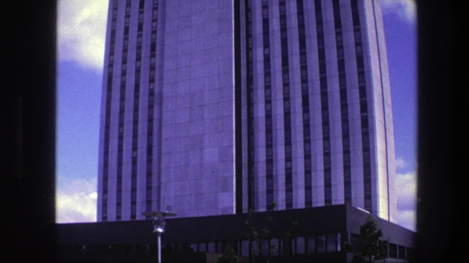 1975 Copenhagen 8mm frame showing the high-rise from street level, with a lamp, trees, and building base below the concrete facade.