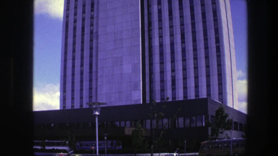 1975 Copenhagen 8mm frame of the high-rise and its street-level base, with lamps, trees, vehicles, and blue sky visible.