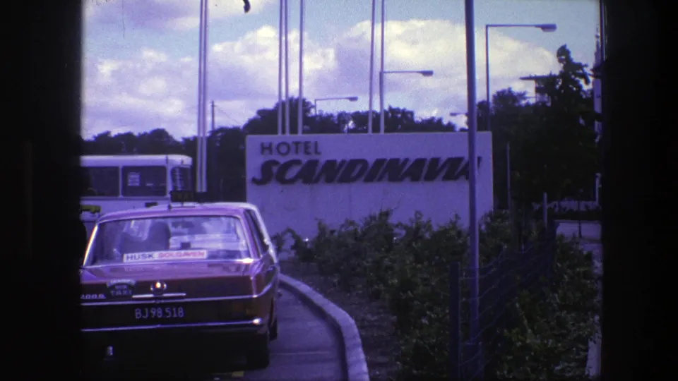 1975 Copenhagen view of the Hotel Scandinavia sign with a taxi in front, road edge, flags or poles, lamps, trees, and a bus behind.