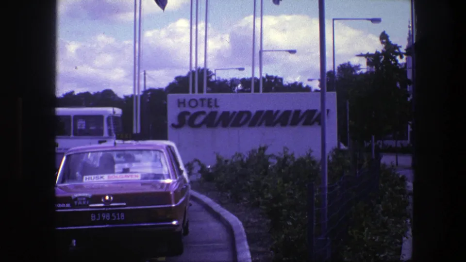 1975 Copenhagen 8mm frame showing the rear of a red taxi near the Hotel Scandinavia sign, with a bus and roadside plantings visible.