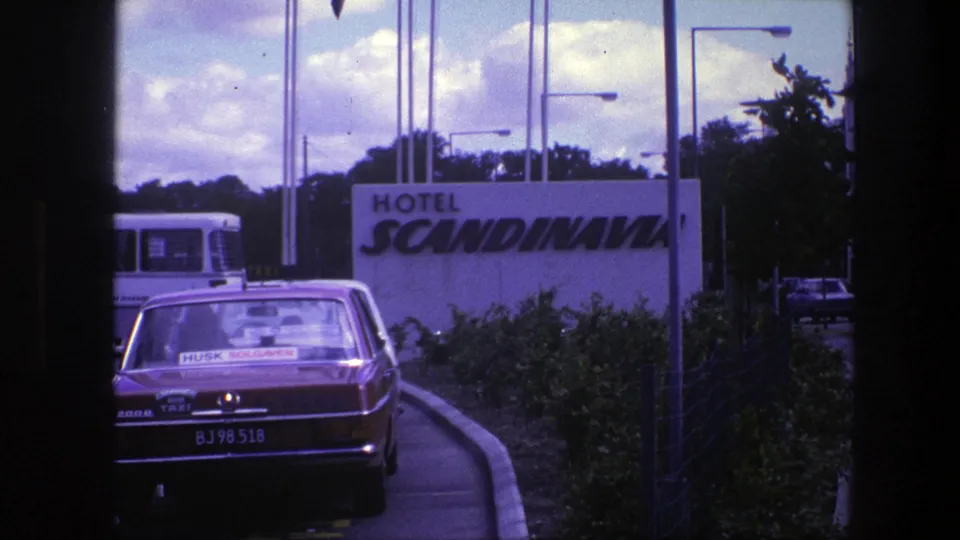 1975 Copenhagen frame with a red taxi, Hotel Scandinavia sign, dark window framing, bus, shrubs, and streetlights in view.