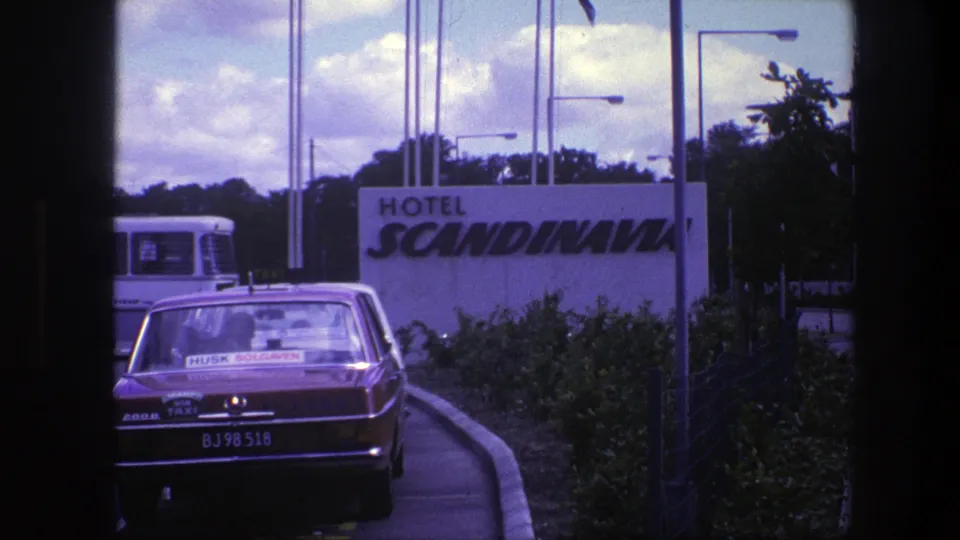 1975 Copenhagen hotel approach with a red taxi in the foreground, the Hotel Scandinavia sign, a white bus, shrubs, and tall poles.