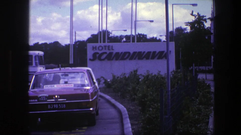 Final 1975 Copenhagen frame showing the parked red taxi at the Hotel Scandinavia driveway, with sign, bus, shrubs, and sky visible.