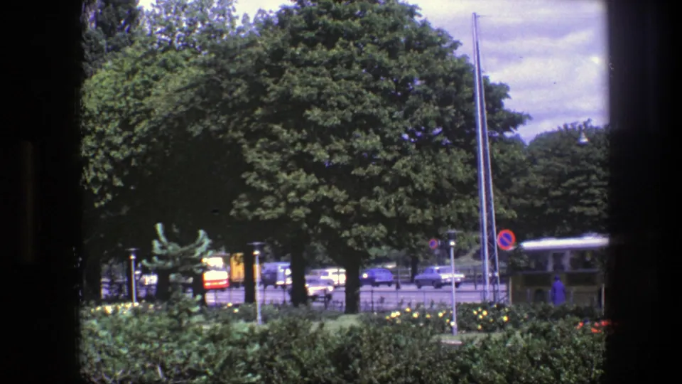 1975 Copenhagen street view showing trees, foreground hedges, tall poles, a no-parking-style sign, cars, and a yellow bus.