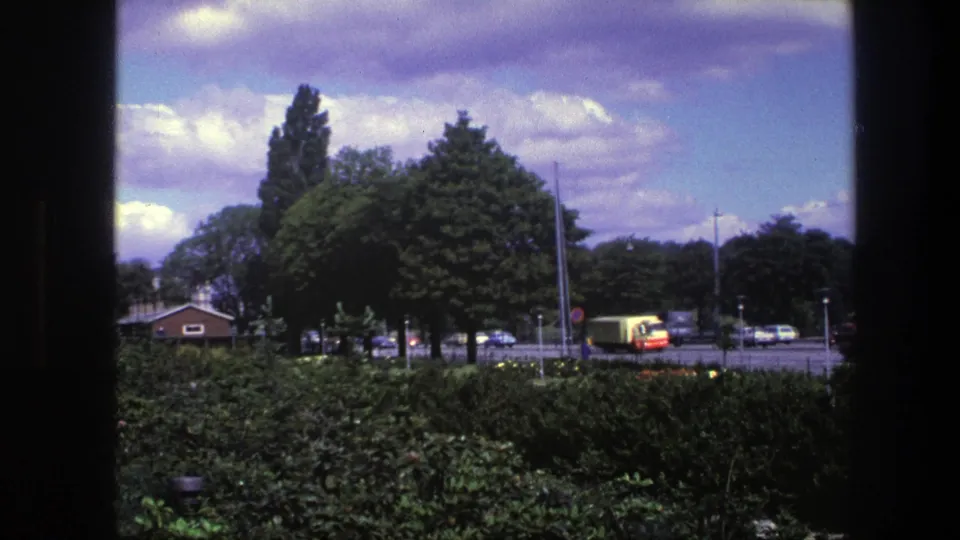 Copenhagen park-edge view in 1975, with hedges, large trees, street traffic, and a yellow truck or bus moving along the road.