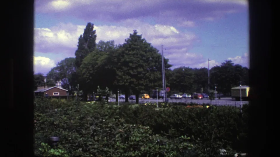 1975 Copenhagen 8mm frame showing dense park planting, a street beyond, cars, and a bus or large vehicle near the right side.