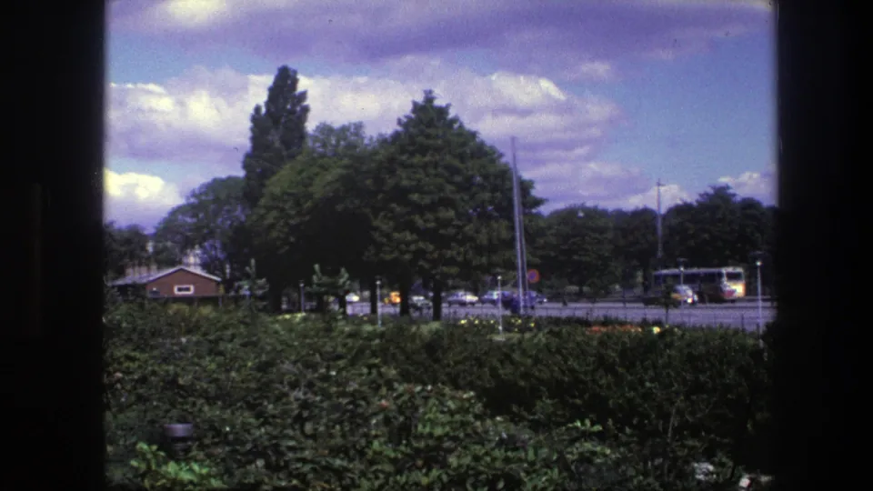 Late clip frame from 1975 Copenhagen with dark film edges around a park-side street view, cars, trees, and a bus or truck at right.