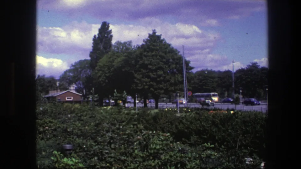Copenhagen urban park and street in 1975, with dense greenery, a small brick building, cars, and a yellow bus in the distance.