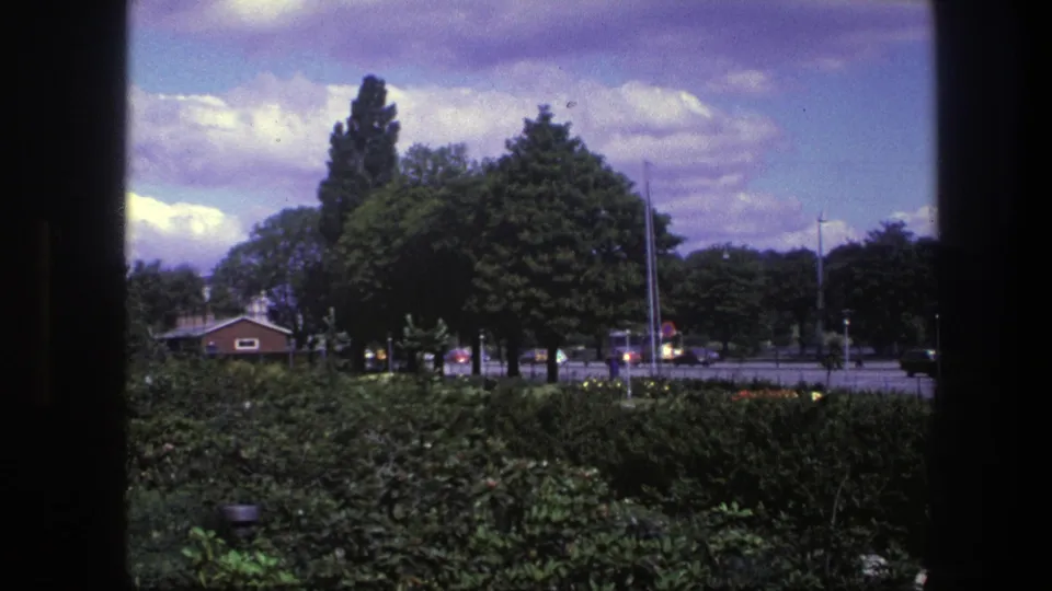 1975 Copenhagen 8mm frame showing a green park edge, cloudy sky, street lamps, cars, and a small brick building.