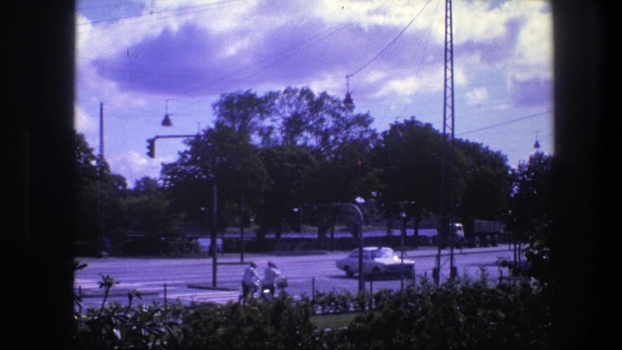 1975 Copenhagen street view filmed from behind a dark frame, with cyclists crossing, cars on the road, trees, wires, and cloudy sky.