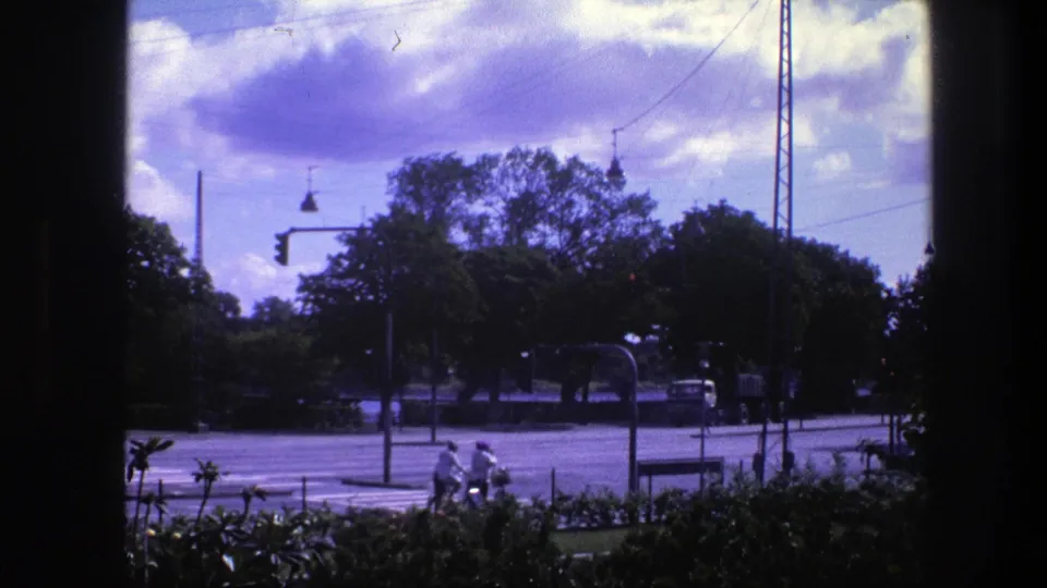 Copenhagen road traffic in 1975, seen through a dark vertical frame with two cyclists, a car, a truck, trees, and overhead street wires.