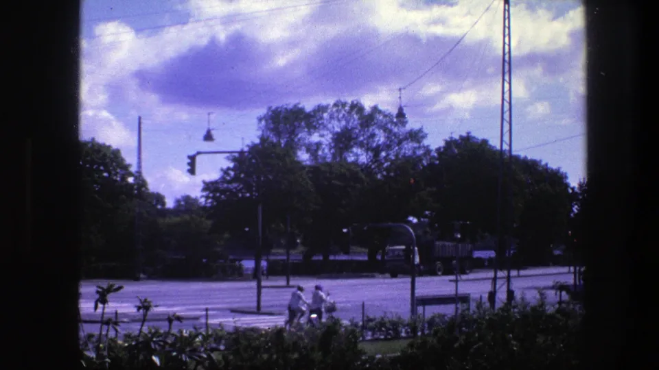 1975 Copenhagen traffic view with two cyclists in front, a large vehicle on the road, overhead wires, trees, and strong 8mm color shift.