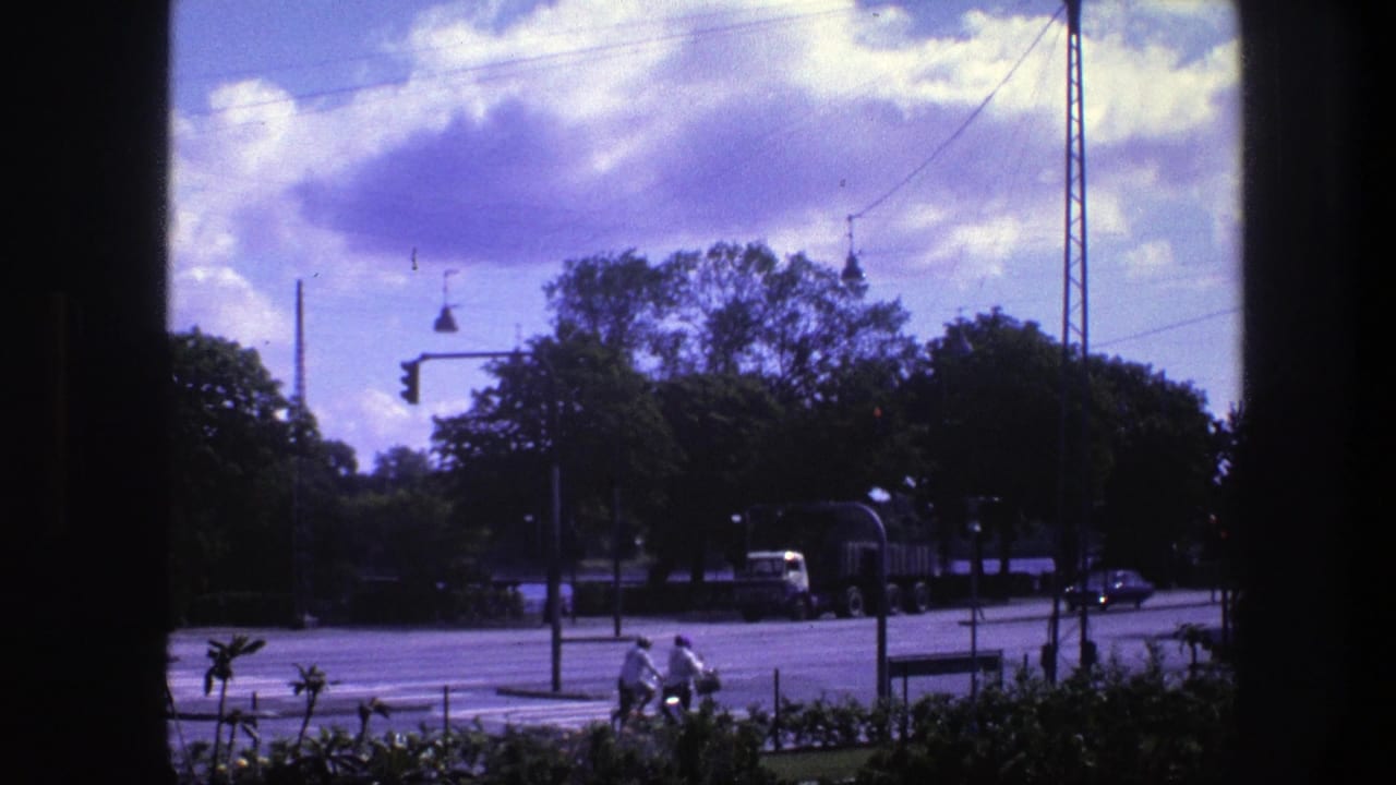 1975 Copenhagen street view from a window, showing cyclists near a crosswalk, traffic lights, trees, and a truck beneath purple-tinged clouds.