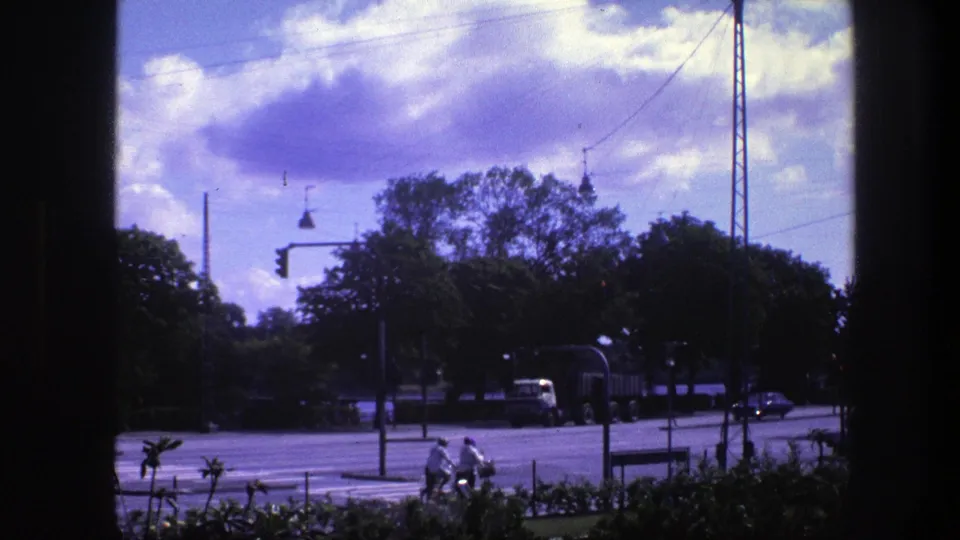 1975 Copenhagen street view from a window, showing cyclists near a crosswalk, traffic lights, trees, and a truck beneath purple-tinged clouds.