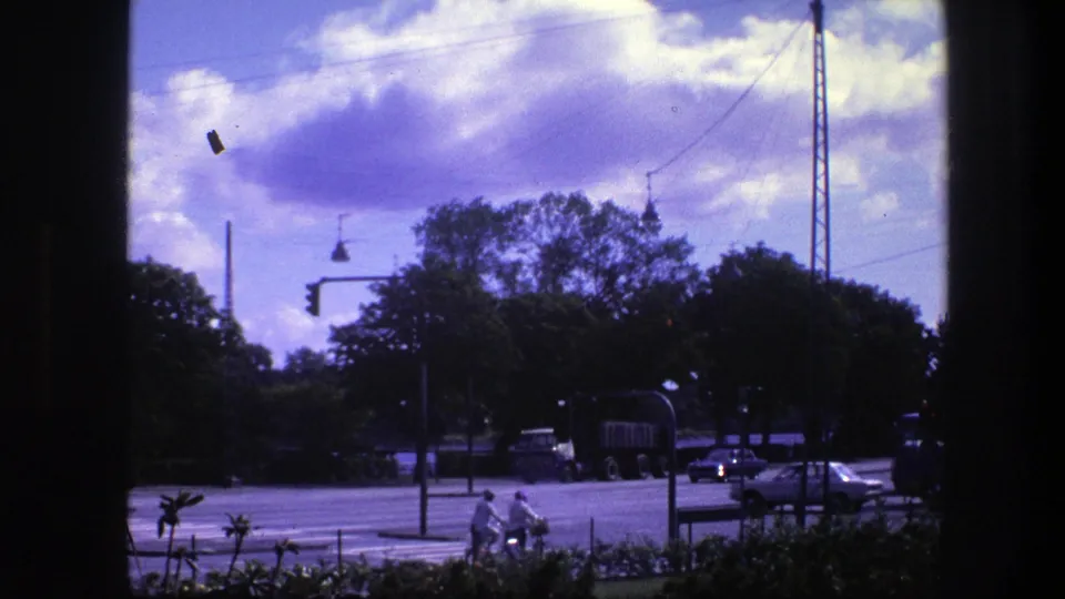 Vintage 8mm view of a Copenhagen intersection with cyclists, a large truck, a passing car, traffic lights, trees, and dark window framing.