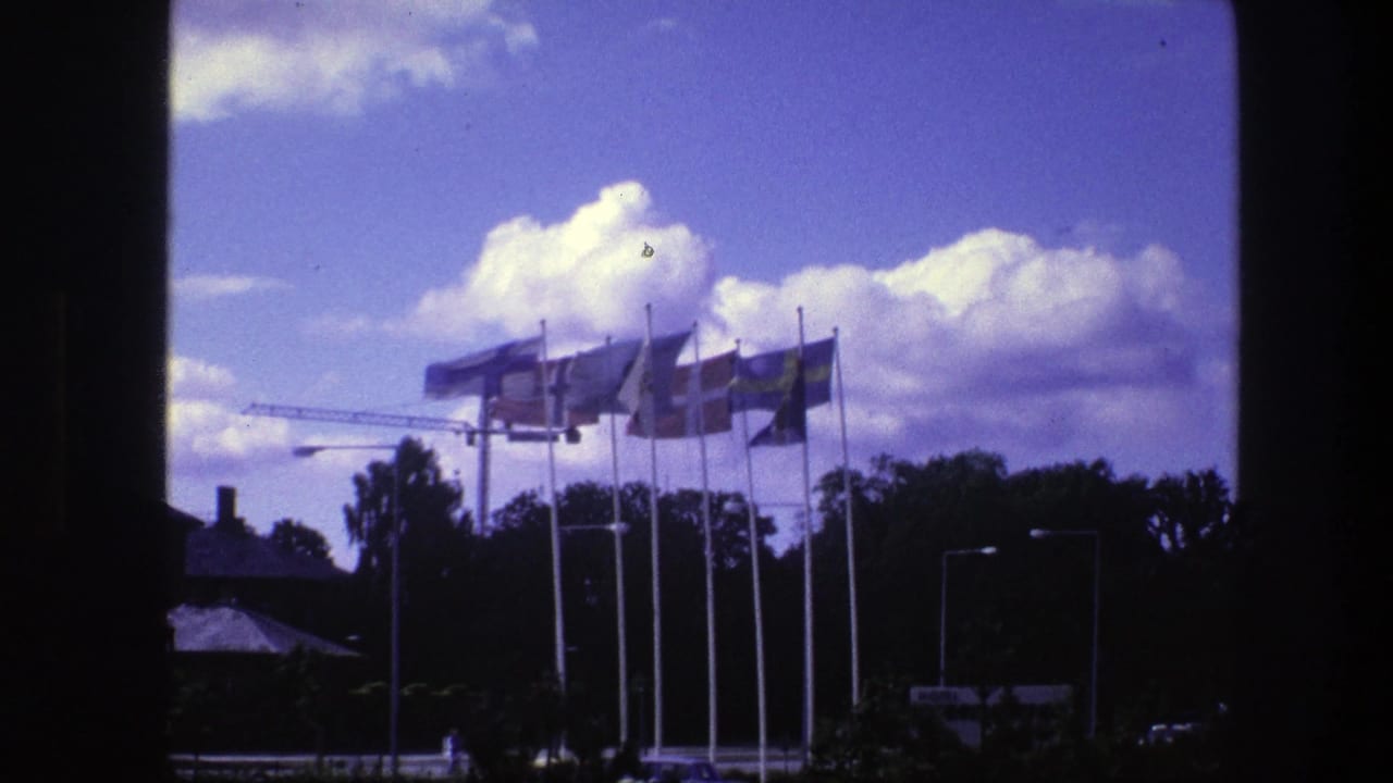 1975 Copenhagen 8mm frame showing international flags waving near a construction crane, viewed through a dark window opening.