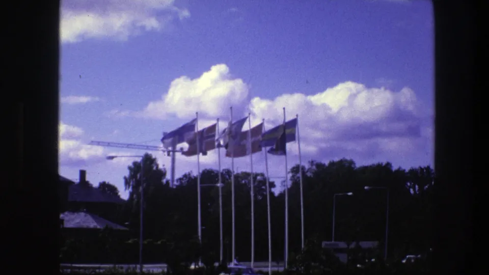 1975 Copenhagen 8mm frame holding on international flags, with bright clouds, street lamps, and shadowed framing from a window or opening.