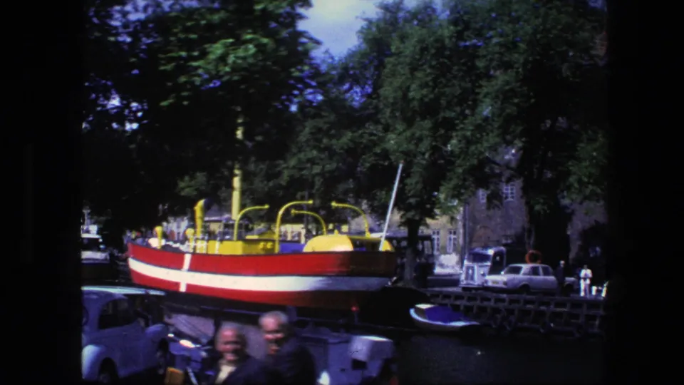 Copenhagen canal-side 8mm frame showing a red-and-white boat, trees, older cars, and blurred adults passing close to the camera.