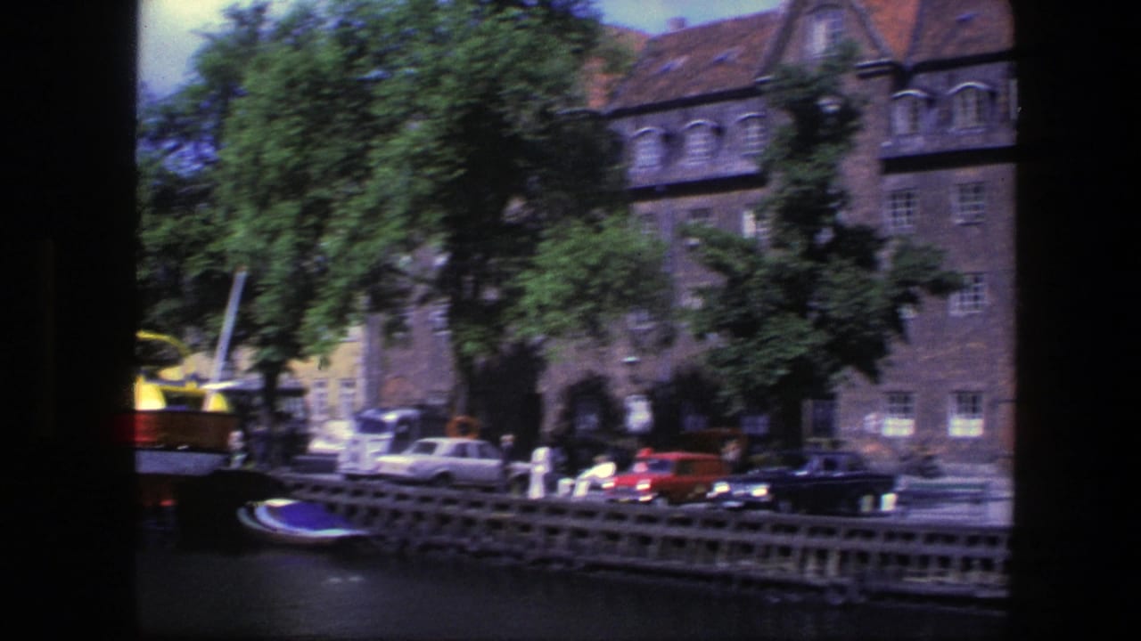 Canal view with historic brick buildings, trees, parked cars, pedestrians, and a small boat at the left edge.