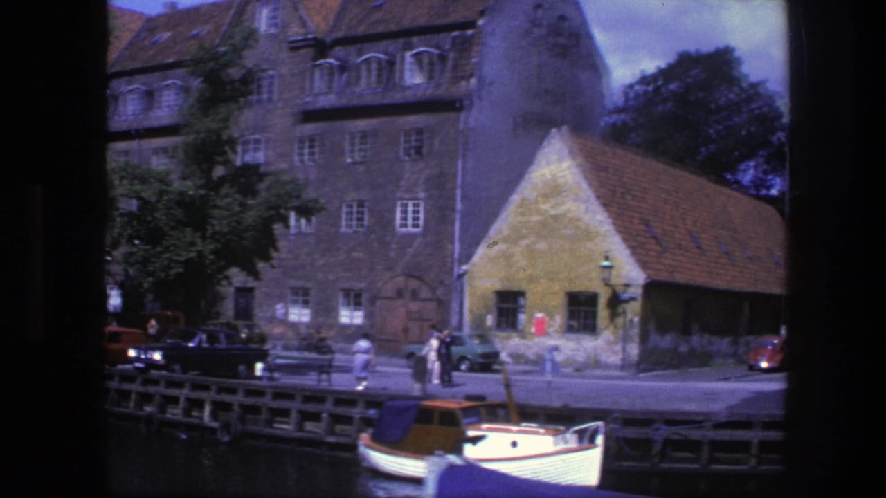 Waterfront buildings, a red-roofed structure, pedestrians, parked cars, and a small boat on the canal.