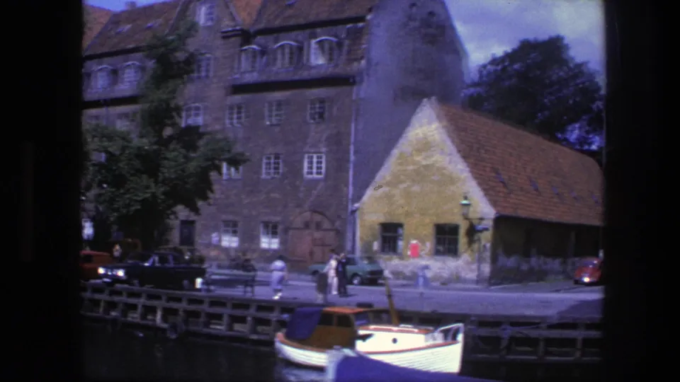 1975 Copenhagen waterfront with historic buildings, a smaller red-roofed structure, distant pedestrians, parked cars, and a white boat on the canal.