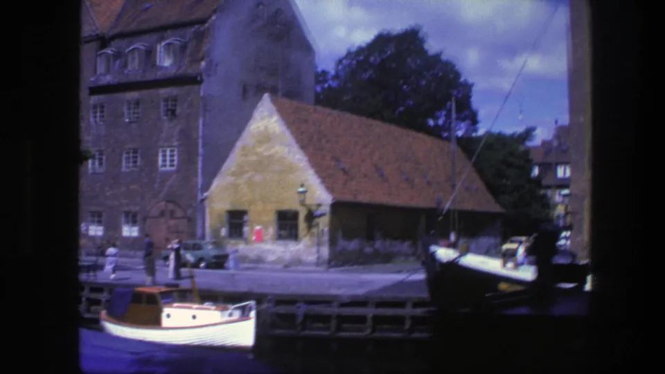 1975 Copenhagen canal view centered on a red-roofed waterfront building, distant pedestrians, parked cars, and a small white boat in the foreground.