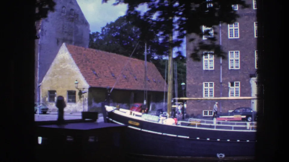 1975 Copenhagen canal frame with a moored dark-hulled boat, red-roofed waterfront structure, brick building facade, trees, and parked cars.