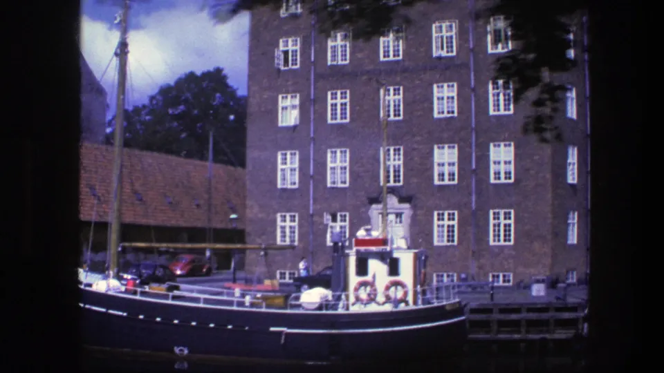 1975 Copenhagen canal scene with a moored boat, mast, brick facade, red-roofed side building, quay, and parked cars.