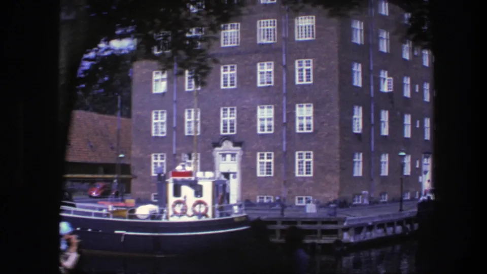 Copenhagen canal view with the boat stern, life rings, brick building, quay edge, parked car, and small pedestrians near the water.