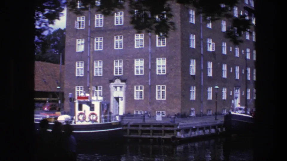 1975 Copenhagen canal view with a brick building, white-framed windows, wooden dock, and small boat moored in front.