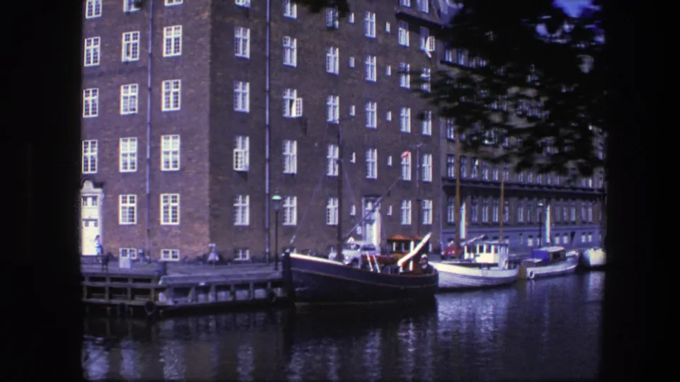 1975 Copenhagen waterfront view with a brick building corner, white windows, dock structure, and a dark moored boat.