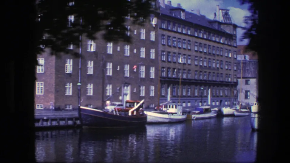 1975 Copenhagen canal view with brick waterfront buildings, several moored boats, reflected water, and a blurred passerby near the frame edge.