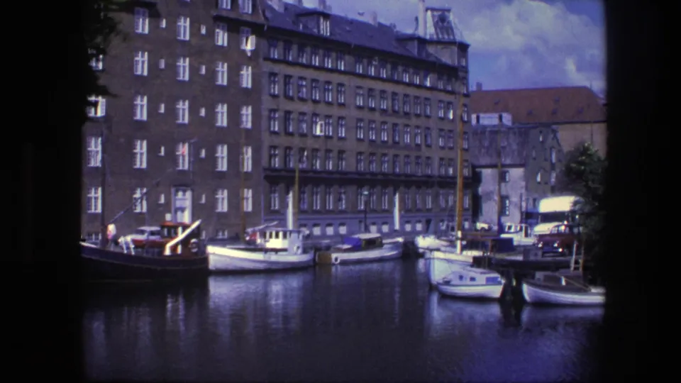 Copenhagen waterfront buildings and moored small boats appear across calm canal water in faded 8mm footage from 1975.
