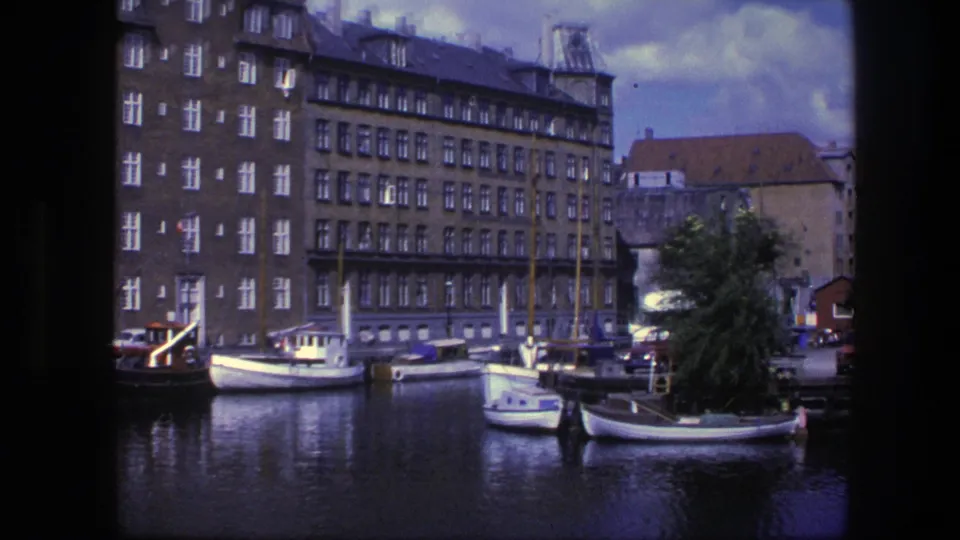 1975 Copenhagen canal scene with moored boats, brick housing, parked cars, and a tree at the quay in home-movie footage.