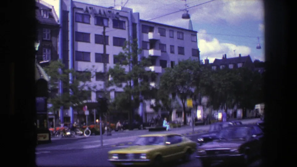 1975 Copenhagen street frame with motion-blurred cars crossing the foreground, pedestrians, trees, and commercial buildings along the avenue.