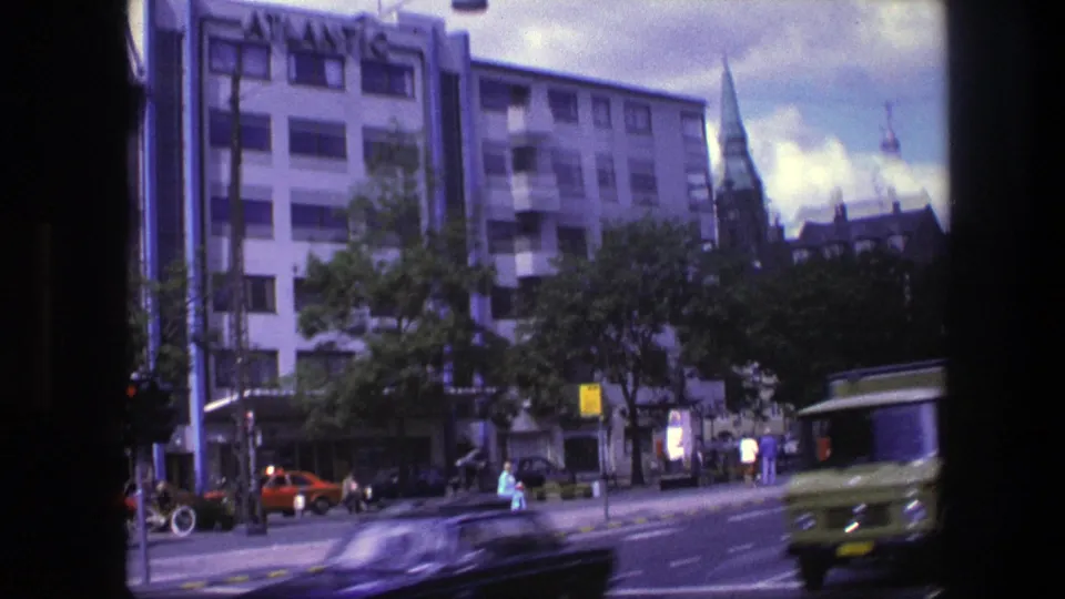 Late-frame Copenhagen avenue view from 1975 with passing vehicles, pedestrians near the crossing, trees, buildings, and a distant spire.