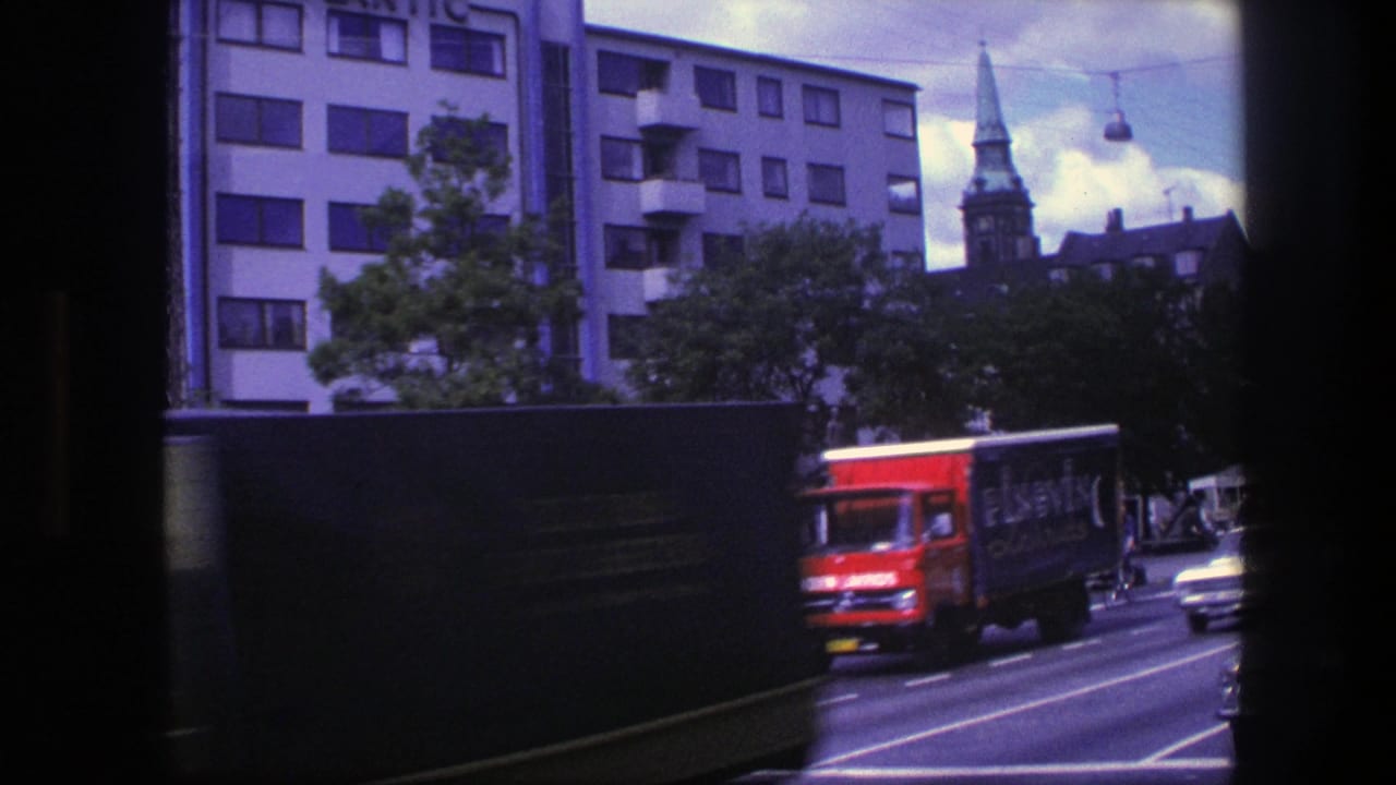 1975 Copenhagen street view from moving 8mm footage, with traffic, apartment blocks, overhead cables, and a church steeple.