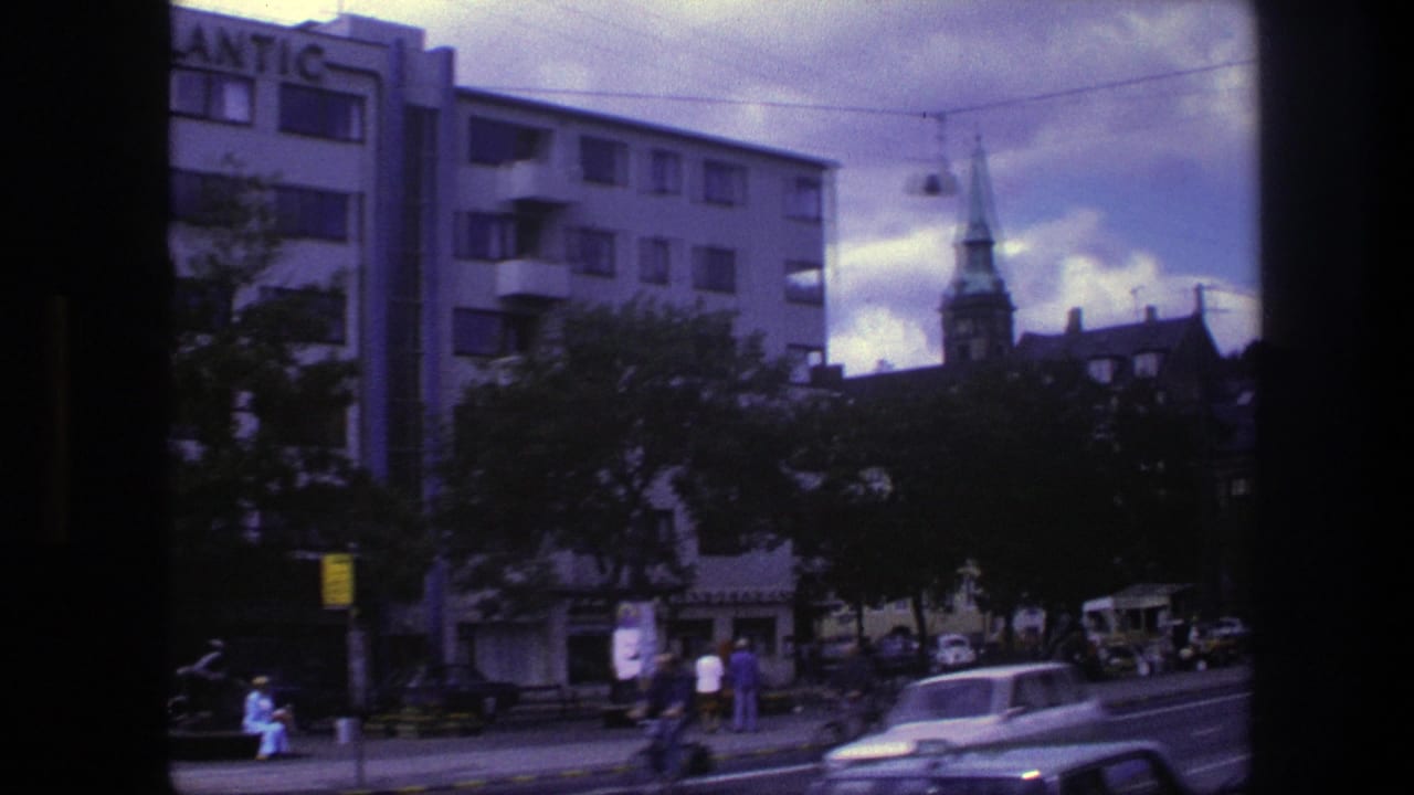 Street scene with apartment facade, church steeple, parked cars, pedestrians, trees, and overhead cables.