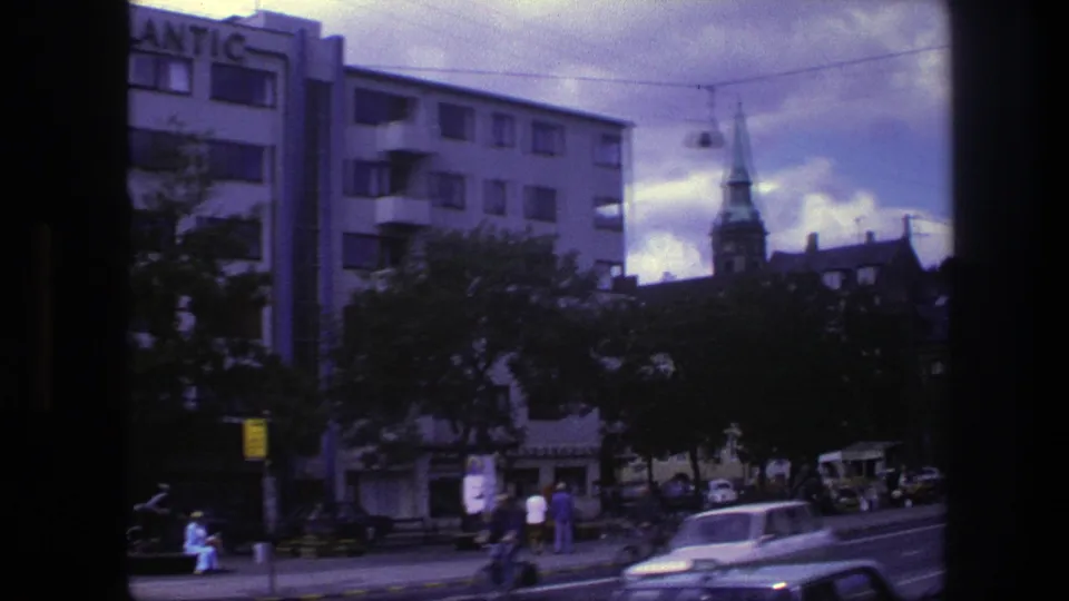 1975 Copenhagen street scene with pedestrians, older cars, trees, apartment blocks, and a church steeple beyond the road.