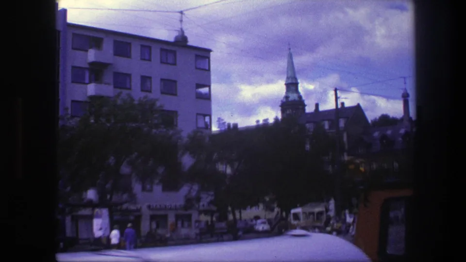 1975 Copenhagen view past trees and pedestrians toward an apartment block and church steeple in soft 8mm color.