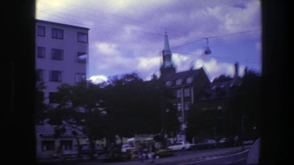 1975 Copenhagen street view with church steeple, apartment buildings, passing cars, pedestrians, and overhead street cables.