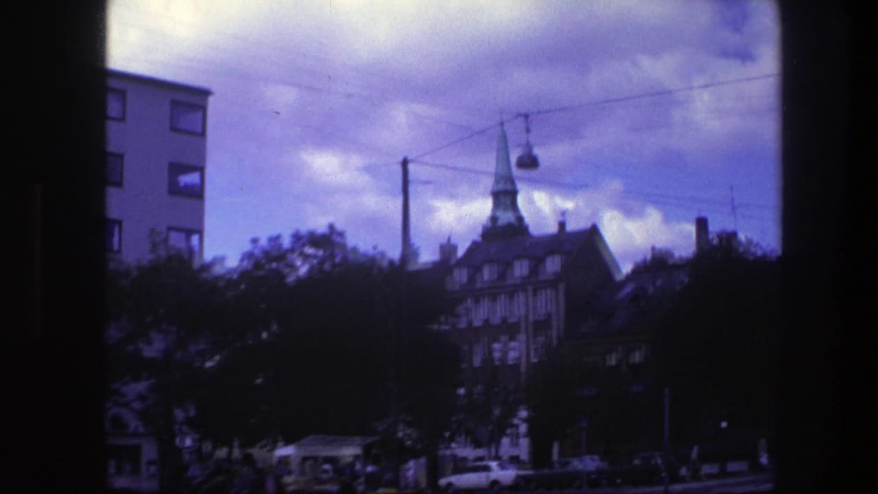 Copenhagen street view with church spire, trees, cars, overhead wires, and cloudy blue sky.