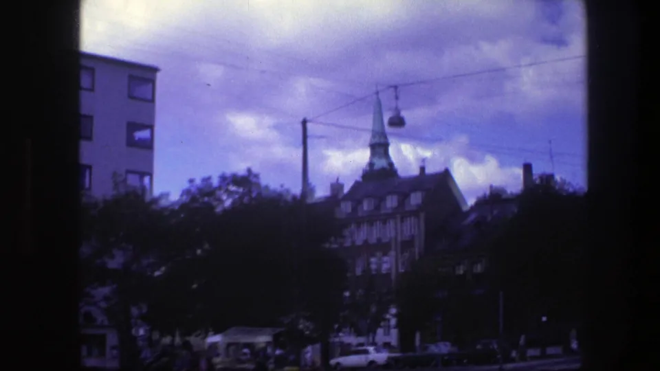 1975 Copenhagen street view from a moving vehicle, with a green church spire, roadside trees, cars, overhead wires, and bright clouds.