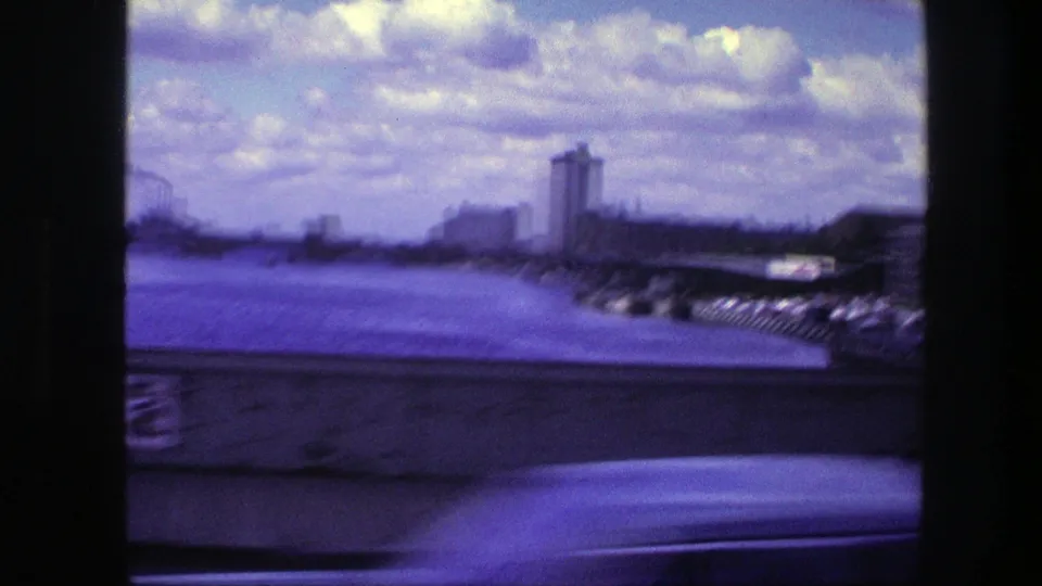 1975 Copenhagen waterfront frame with motion blur, water, a low riverside wall, parked cars, a distant tower, and scattered clouds.