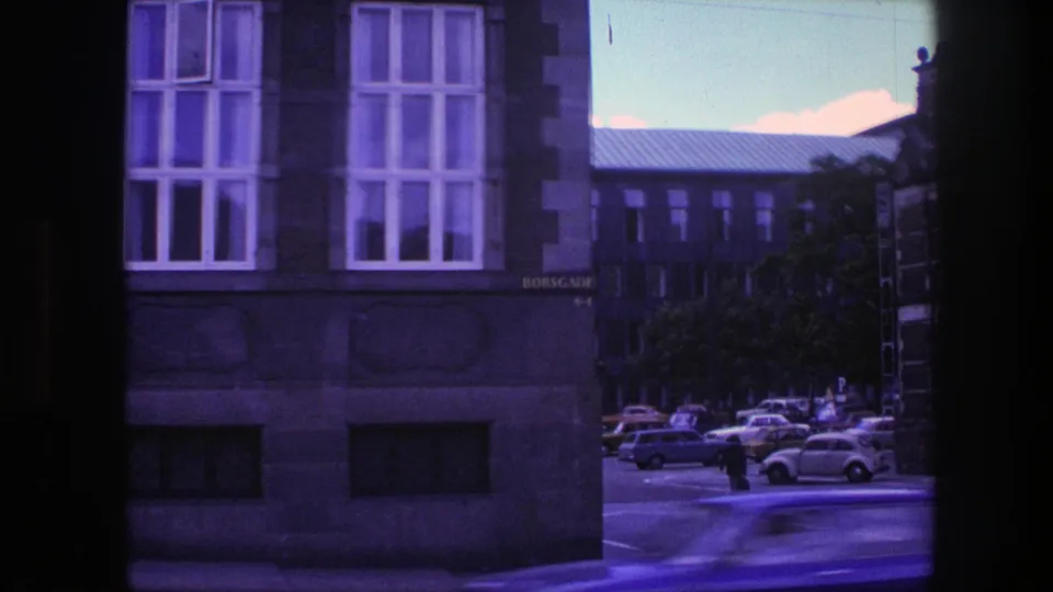 1975 Copenhagen drive-by frame showing a building facade, parked cars, a distant pedestrian, and street markings under a strong blue-purple cast.