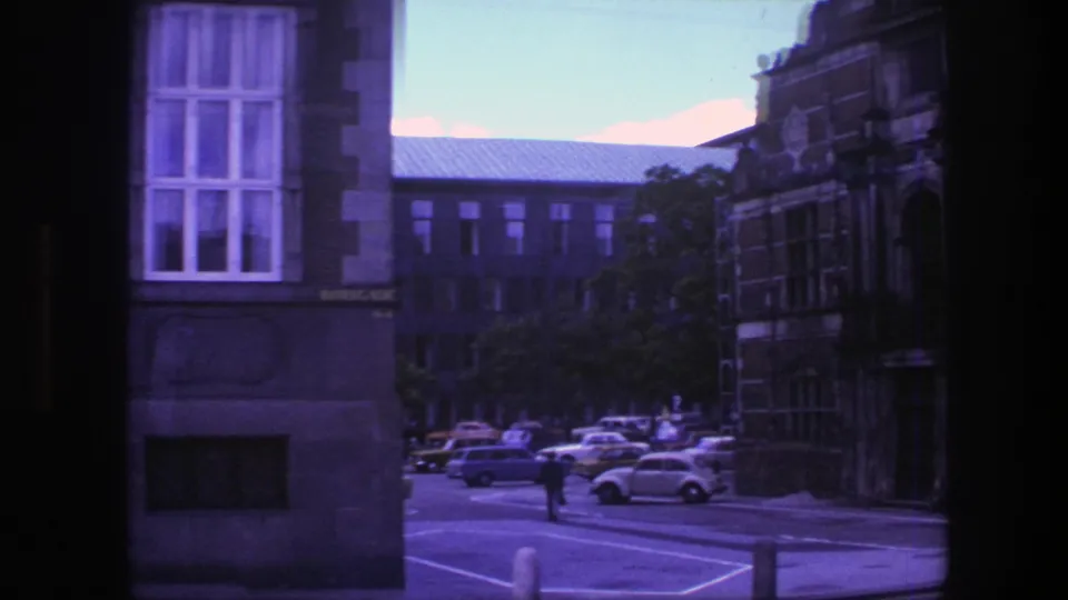 1975 Copenhagen street view with parked cars, a pedestrian, trees, building fronts, and visible road markings from a moving vehicle.